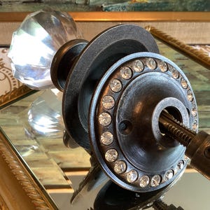 May include: A door knob featuring a clear, faceted glass handle attached to a dark, weathered metal circular plate. The plate is embellished with a ring of small, clear, sparkling crystals set around the central opening and a metal spindle.