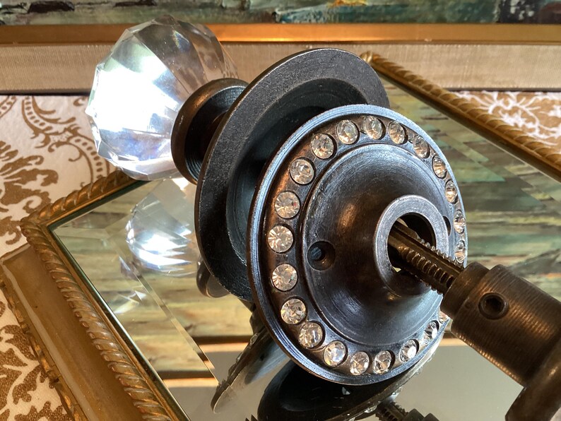May include: A door knob featuring a clear, faceted glass handle attached to a dark, weathered metal circular plate. The plate is embellished with a ring of small, clear, sparkling crystals set around the central opening and a metal spindle.