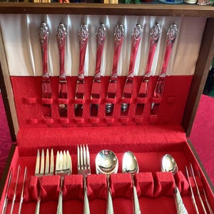 May include: A wooden silverware case with a red velvet interior. The case holds multiple pieces of silverware, including knives, forks, and spoons. The silverware has a decorative floral design on the handles. The case is open, revealing the contents.