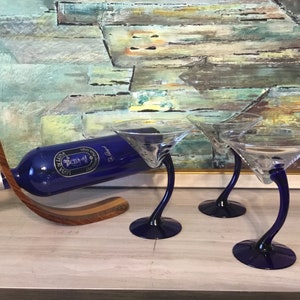 May include: Three blue glass martini glasses with a curved stem and a bottle of blue liqueur on a wooden stand.