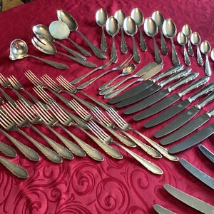 Puede incluir: Un surtido de cubiertos plateados, que incluye tenedores, cucharas, cuchillos y utensilios para servir, dispuestos sobre un mantel rojo. La cubertería tiene mangos decorativos y está preparada para una cena formal.