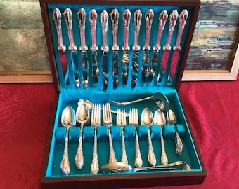 Op de afbeelding: Een zilverkleurige bestekset in een donkerhouten opbergdoos met een fel turquoise fluwelen binnenkant. De set bevat twaalf messen die rechtop in het deksel staan, samen met diverse lepels, vorken en een soeplepel in de onderkant van de doos.
