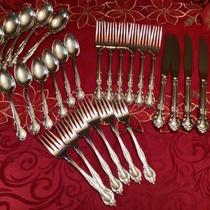 May include: A set of silverware with a silver finish. The set includes spoons, forks, and knives. The silverware is arranged on a red tablecloth with a floral pattern.