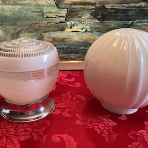 May include: Two vintage white glass light fixtures. One has a clear, ridged top and a decorative band. The other is a round, ribbed globe. Both sit on a red floral tablecloth, with a framed painting in the background.