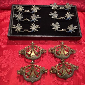 Op de afbeelding: Een set van tien antieke messing kastbeslag trekt. De trekt zijn in twee stijlen, met sierlijke bloemmotieven. De trekt worden getoond op een zwarte fluwelen achtergrond.