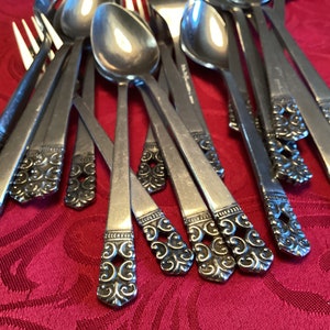 May include: A close-up of a set of silver-plated silverware with ornate handles. The handles are decorated with a swirling pattern and have a slightly raised design. The silverware is arranged in a pile, with the handles facing up.