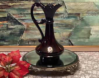 May include: A dark red glass pitcher with a decorative handle and a round base. The pitcher is sitting on a silver mirrored stand with ornate details.