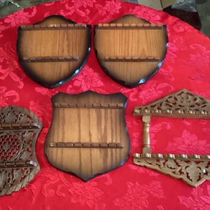 Puede incluir: Una colección de estantes de madera para cucharas en varias formas de escudo y decorativas. Los estantes son de madera con un acabado marrón oscuro y cuentan con múltiples ranuras para sostener cucharas. El fondo es un mantel rojo floral.