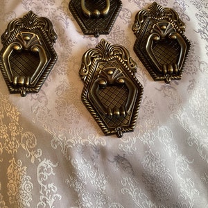 May include: Four ornate gold-toned metal door handles with intricate designs. Each handle features a ring pull and a decorative, textured surface.