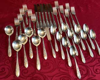 May include: A set of silverware with a floral pattern on the handles. The set includes forks, spoons, and knives. The silverware is arranged in rows on a red tablecloth.