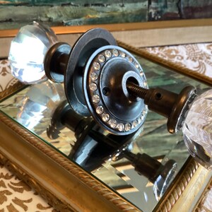 May include: A door handle set featuring two clear, multifaceted glass knobs attached to a dark metal spindle. The central circular metal plate is decorated with a ring of small, clear embedded rhinestones, providing a contrast to the glass.