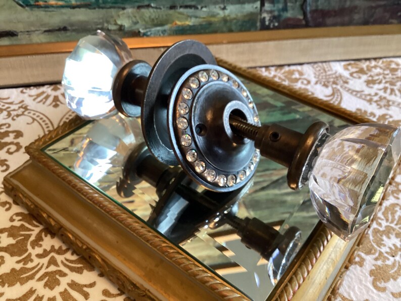 May include: A door handle set featuring two clear, multifaceted glass knobs attached to a dark metal spindle. The central circular metal plate is decorated with a ring of small, clear embedded rhinestones, providing a contrast to the glass.
