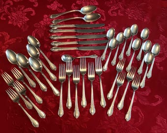 Può includere: Una collezione di posate color argento, tra cui cucchiai, forchette e coltelli, disposte su una tovaglia rossa a motivi floreali. Le posate hanno manici decorativi e una finitura lucida.