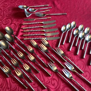 May include: A collection of stainless steel silverware, including spoons, forks, and knives, arranged on a red patterned tablecloth. The silverware has a decorative handle design.
