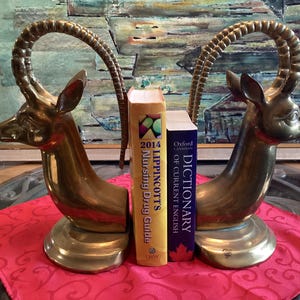 May include: Two brass antelope bookends with curved horns, either side of two books. The bookends have a detailed design and are placed on a red cloth. The books are titled "Lippincott's Nursing Drug Guide" and "Oxford Dictionary of Current English".