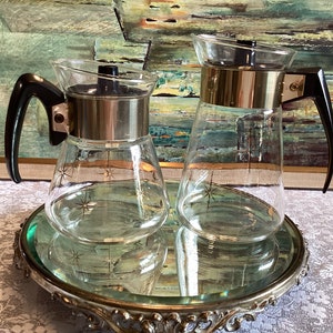 May include: Two vintage glass coffee carafes with black handles and gold accents. The carafes have a starburst design and are sitting on a silver mirrored tray.