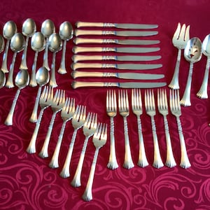 May include: A collection of silver-colored flatware, including spoons, forks, and knives, arranged on a red patterned tablecloth. The silverware has decorative handles and is likely for formal dining.