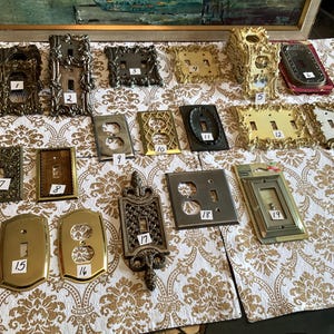 Könnte beinhalten: Eine Sammlung von Vintage-Lichtschalter- und Steckdosenabdeckungen in verschiedenen Stilen und Ausführungen. Die Abdeckungen umfassen verzierte, goldene, bronzefarbene und silberne Designs, wobei einige aufwendige Muster und dekorative Rahmen aufweisen.