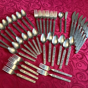 Può includere: Un set completo di posate in acciaio inossidabile, tra cui cucchiai, forchette, coltelli e coltelli da burro, disposte su una tovaglia rossa fantasia. Le posate presentano manici ornati e incisi.