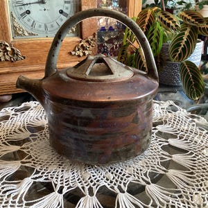 May include: A round ceramic teapot with a mottled brown and dark blue glaze. It features a high arched handle, a short spout, and a lid with a geometric cutout design. The surface has a textured, earthy appearance with visible colour variations.