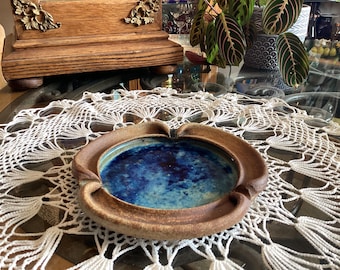 Op de afbeelding: Een ronde keramische asbak met een textuurrijke, ongeglazuurde bruine buitenrand en een gladde, glanzende binnenkant met een levendige mix van diepblauw en turquoise glazuur. Het object heeft vier duidelijke inkepingen in de rand.