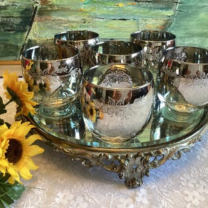 May include: Six silver-plated drinking glasses with floral etched designs are arranged on a mirrored tray with an ornate gold-coloured base. The glasses reflect the surrounding environment. Yellow sunflowers are in the foreground.