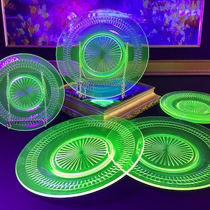 May include: A collection of vintage green glass plates, including dinner plates and smaller plates. The plates have a decorative etched design and are arranged on a dark surface. The plates are illuminated with a bright green glow.