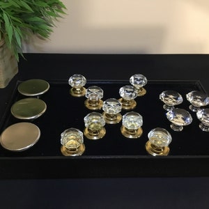 Peut inclure: Une collection de 13 boutons de porte en verre transparent avec des bases en métal doré, présentés sur un plateau en velours noir. Trois bases en métal doré sont également incluses.