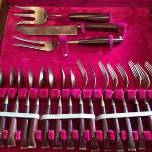 Peut inclure: Un ensemble de 24 couverts en laiton et en bois dans un coffret en bois avec un revêtement en velours rouge. L'ensemble comprend des fourchettes, des couteaux et des cuillères.