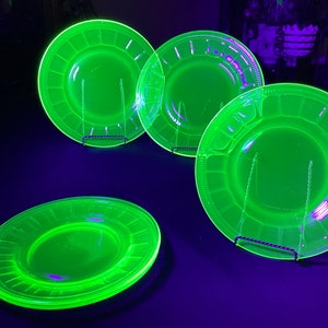 May include: Four green uranium glass plates with a decorative rim. The plates are on black stands and are illuminated with a black light.