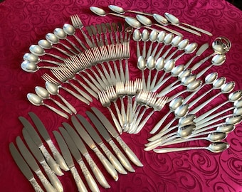 May include: A collection of silver-colored flatware, including spoons, forks, knives, and a ladle, arranged on a patterned red tablecloth. The silverware has decorative handles and is neatly organized, suggesting a formal dining setting.