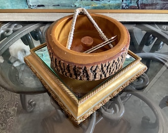 Op de afbeelding: Een ronde houten notenkom met een natuurlijke schors aan de buitenkant en een gladde afwerking aan de binnenkant. In de kom ligt een zilverkleurige metalen notenkraker. De kom staat op een vierkante, goudkleurige voet met een spiegeloppervlak.