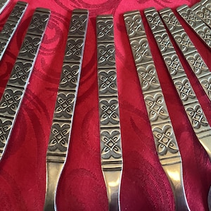 May include: A set of silver-colored flatware with a decorative pattern of repeating geometric shapes. The flatware is arranged in a fan shape on a red tablecloth.