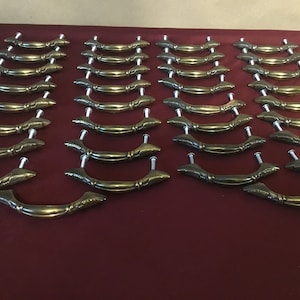 May include: A set of 24 gold-toned cabinet drawer pulls with a decorative design. The pulls have a curved shape and are made of metal.