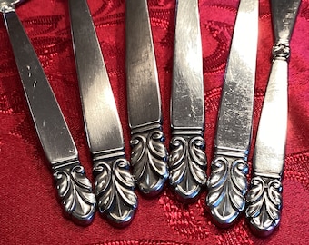 May include: Close-up of six silver flatware pieces with ornate, leaf-shaped handles. The handles are polished and reflect light.