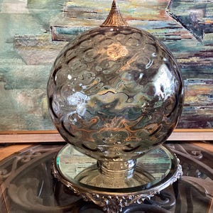 May include: A decorative glass orb with a textured surface, resting on a silver-toned base. The orb has a pointed gold-coloured top. The background features a framed painting with blue, green, and gold tones. The orb is displayed on a glass table.