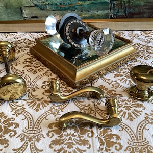 May include: A collection of door hardware including two brass door knobs, two brass lever handles, and one glass door knob set featuring a dark metal base with a ring of clear crystals. The items are arranged on a gold-toned mirrored tray.