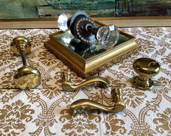 Op de afbeelding: Een verzameling deurbeslag bestaande uit twee messing deurknoppen, twee messing deurkrukken en een glazen deurknopset met een donkere metalen basis en een ring van heldere kristallen, geplaatst op een goudkleurig spiegelplateau.