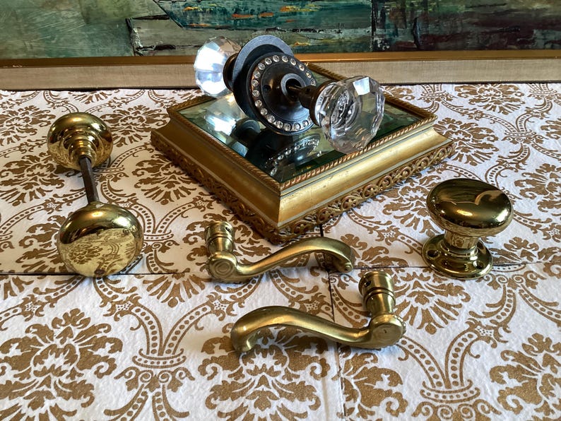 May include: A collection of door hardware including two brass door knobs, two brass lever handles, and one glass door knob set featuring a dark metal base with a ring of clear crystals. The items are arranged on a gold-toned mirrored tray.
