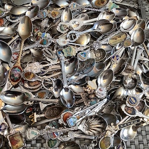 May include: A pile of vintage spoons, mostly silver-coloured metal, with decorative handles and ornate designs. Many spoons have souvenir-style crests and enamel details. The collection includes spoons of various sizes and styles, with detailed engravings.