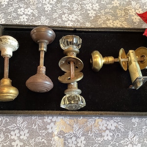 Puede incluir: Cinco pomos de puerta antiguos en una caja forrada de terciopelo negro. Los pomos están hechos de latón, vidrio y madera. Los pomos son de varios estilos y acabados.