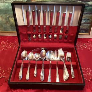 May include: A vintage silverware set in a black wooden case with a red velvet interior. The set includes knives, forks, spoons, and serving utensils. The silverware has a shiny, silver finish, and the case is open, displaying the arrangement.