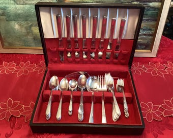 Op de afbeelding: Een vintage bestekset in een zwarte houten kist met een rode fluwelen binnenkant. De set bevat messen, vorken, lepels en serveerbestek. Het bestek heeft een glanzende, zilveren afwerking en de kist is open.