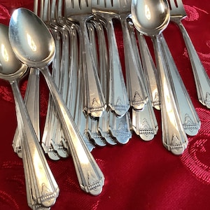 May include: A collection of silver-colored spoons and forks arranged on a red patterned tablecloth. The silverware has a decorative design on the handles. The spoons are in the foreground, with the forks behind them.