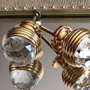 Può includere: Due pomelli per armadi in tono dorato con accenti di cristallo sfaccettato. I pomelli hanno un design decorativo a coste attorno alla base.