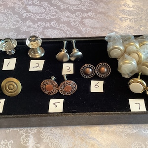 May include: Assortment of 10 cabinet knobs in various styles and finishes, including glass, metal, and ceramic. The knobs are displayed on a black tray with numbers 1 through 7.