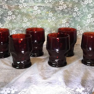 May include: A set of six vintage ruby red drinking glasses. Each glass features a textured, faceted design and a thick base. The glasses are displayed on a white lace tablecloth, with a floral patterned backdrop.