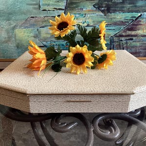 May include: A hexagonal jewellery box with a textured cream and brown speckled finish and a thin gold trim. The lid features a bouquet of artificial yellow sunflowers with dark brown centres and green leaves resting on the flat top surface.