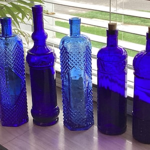 May include: Five cobalt blue glass bottles of varying shapes and designs. The bottles are displayed on a windowsill, with a plant in a white pot visible in the background. The bottles include diamond and textured patterns.