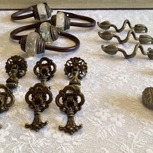May include: An assortment of antique bronze hardware, including curtain tiebacks with clear glass accents, leaf-shaped pulls, and ornate key-shaped drawer pulls. The hardware is arranged on a white floral patterned surface.